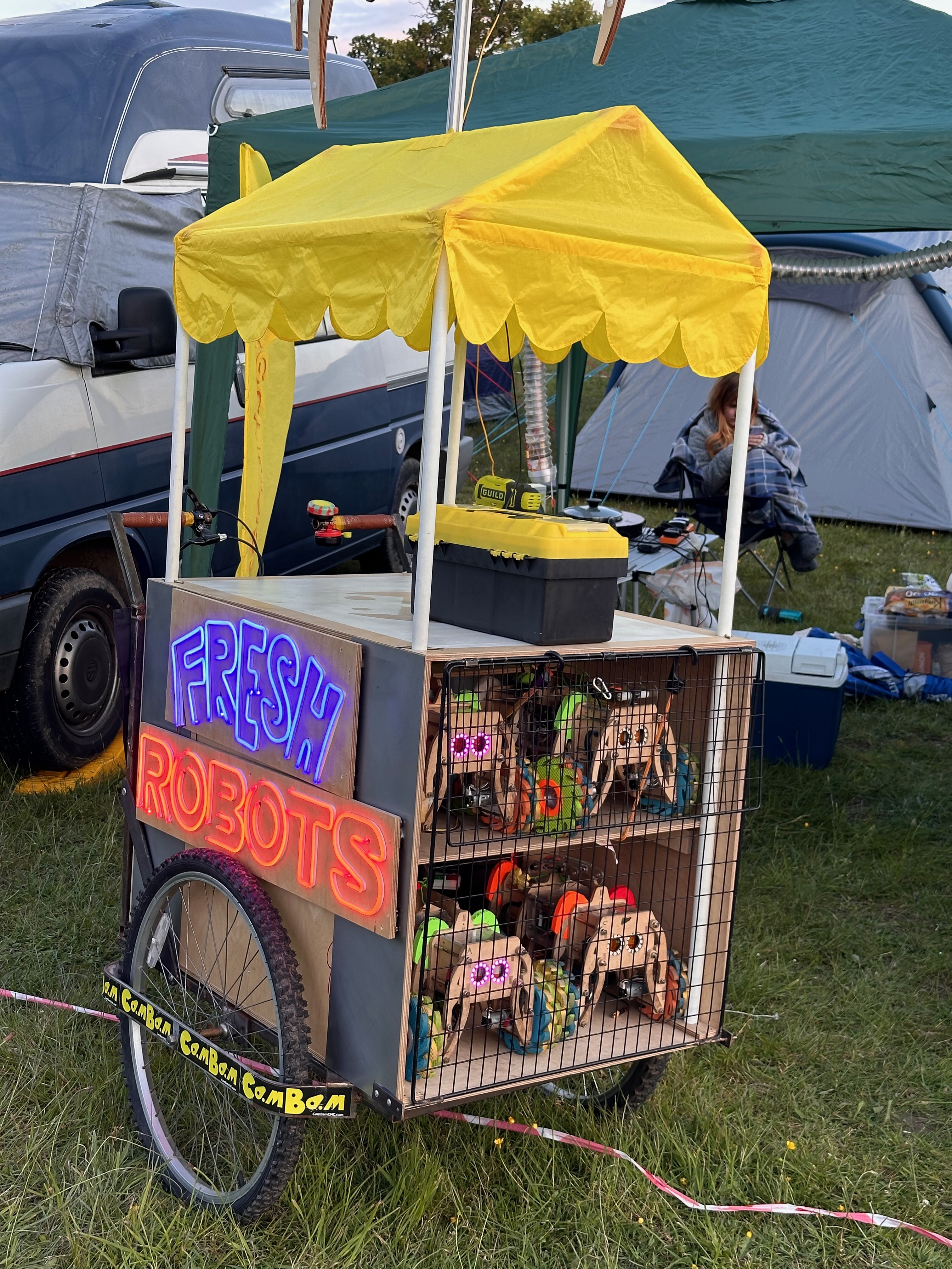 An icecream cart. The side of the cart that would face away from the vendor is lit up with neon lights; 'Fresh Robots'. Tucked inside the cart are two shelves of robots, held in place by a wire mesh. Their eyes look outwards as if they are tyring to escape.