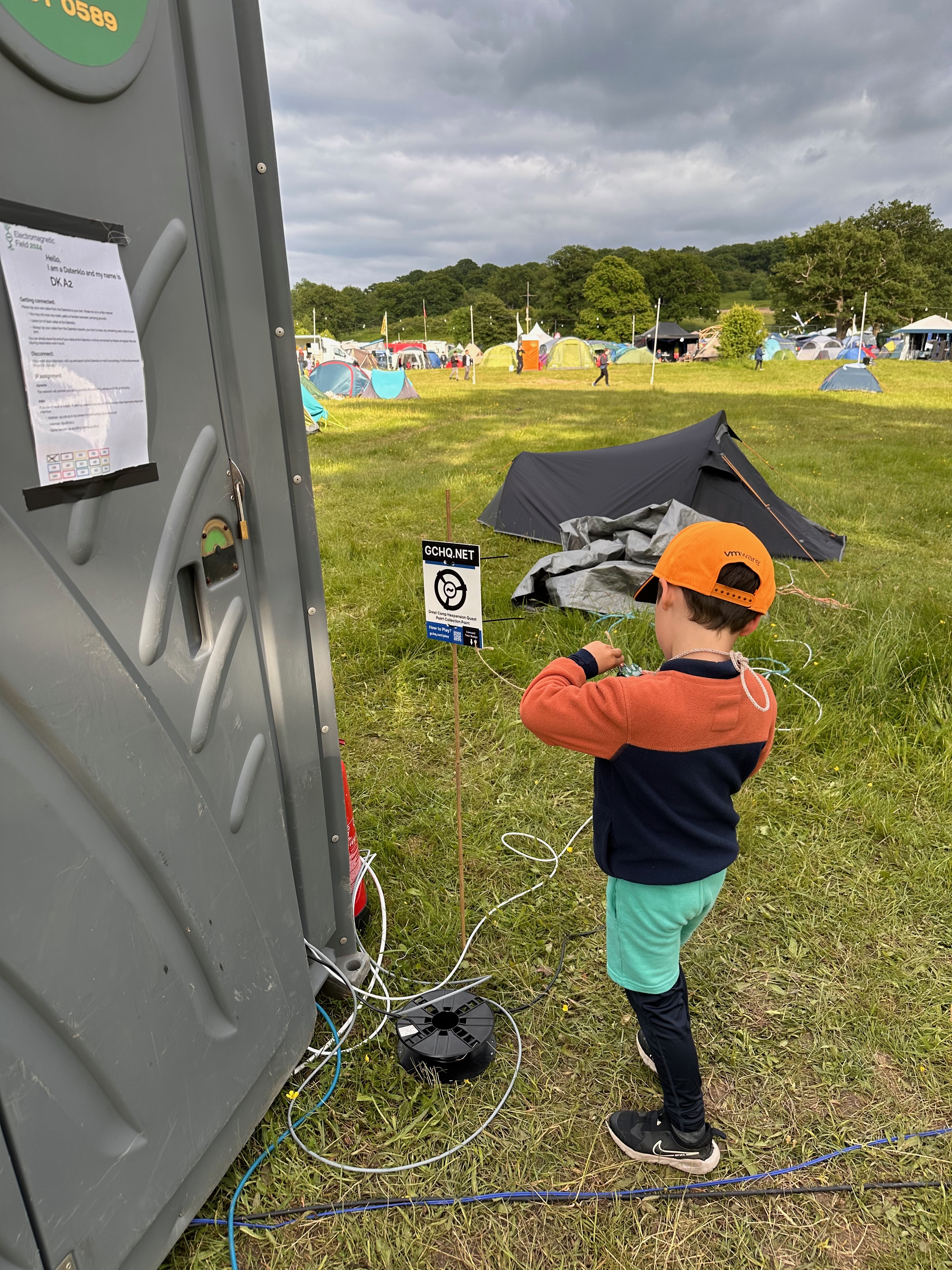 A young boy stands in front of a port-a-loo, facing away from the camera. We can't see what he's doing, but he's plugging something in to a badge held in his hand.
