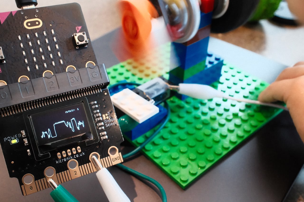 A photograph showing a micro:bit controller with a mini screen. On the screen is a graph. In the background a lego model. Blurred image indicates the model is moving
