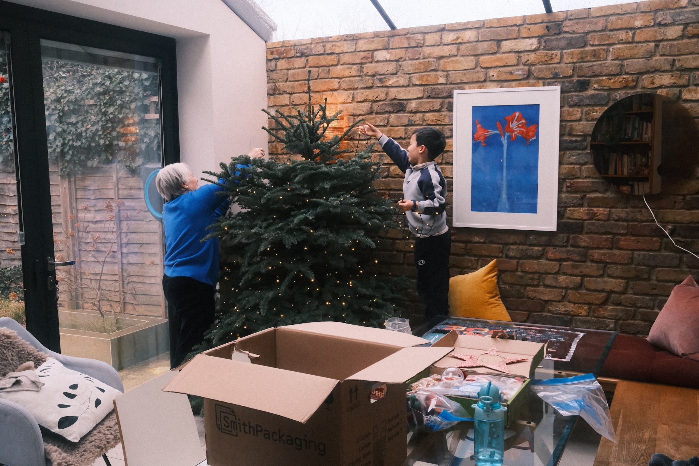 Two people remove the lights from a Christmas tree. In the foreground a cardboard box of decorations sits on a glass table.