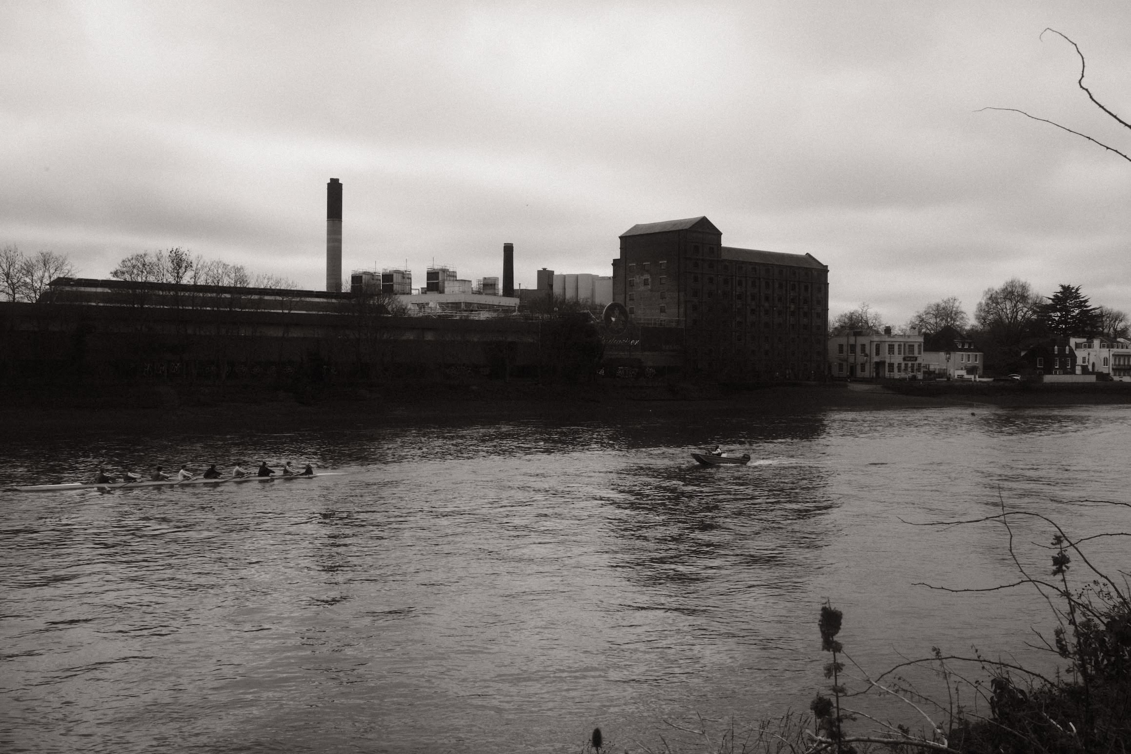 A black and white photo looking out over a river. A rowing boat and safety boat occupy the middle of the river. On the far bank an industrial building and chimney stack mark the skyline.