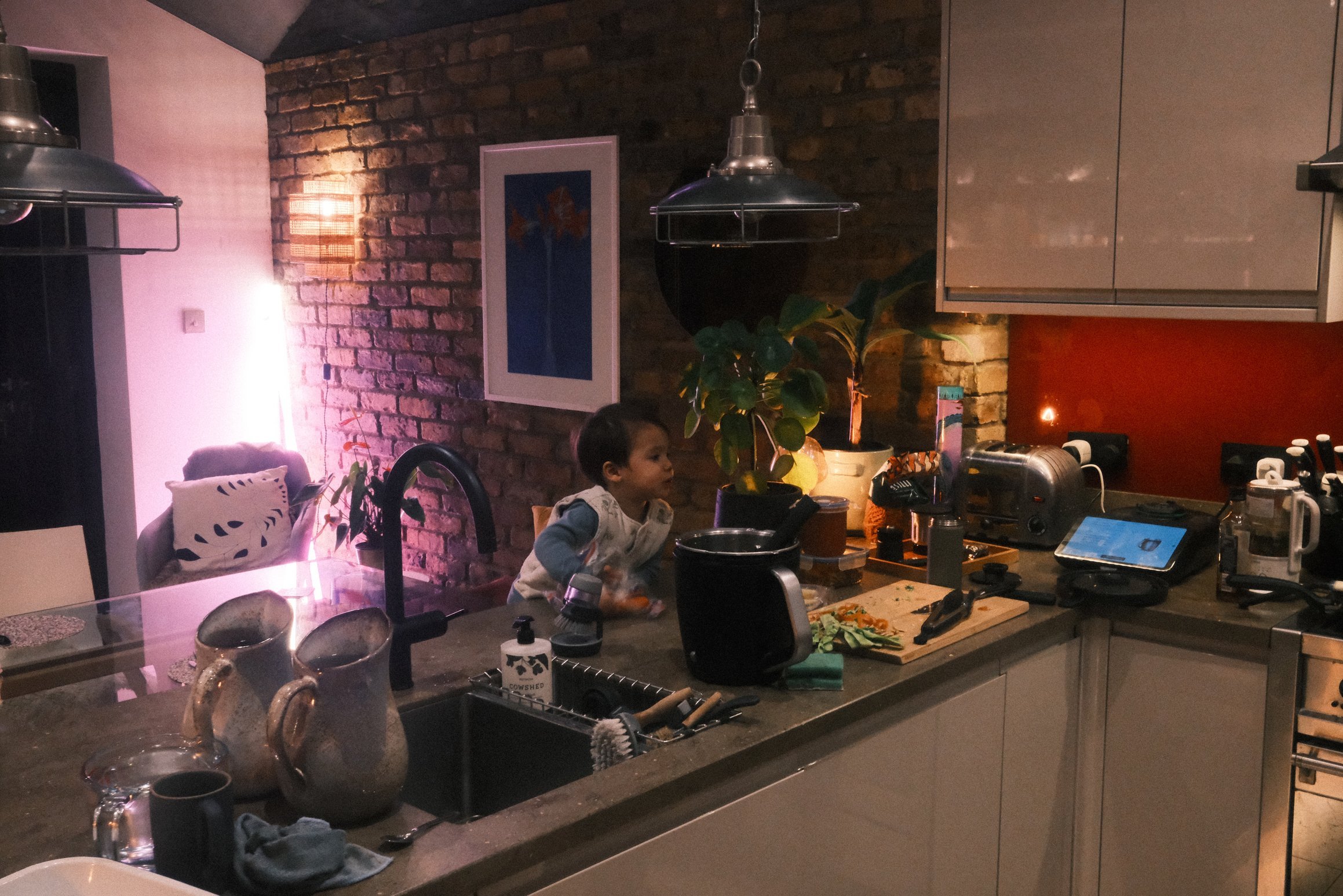 a young boy looks expectantly towards someone or something outside the frame of the image, a busy kitchen worktop indicates meal preparation in progress