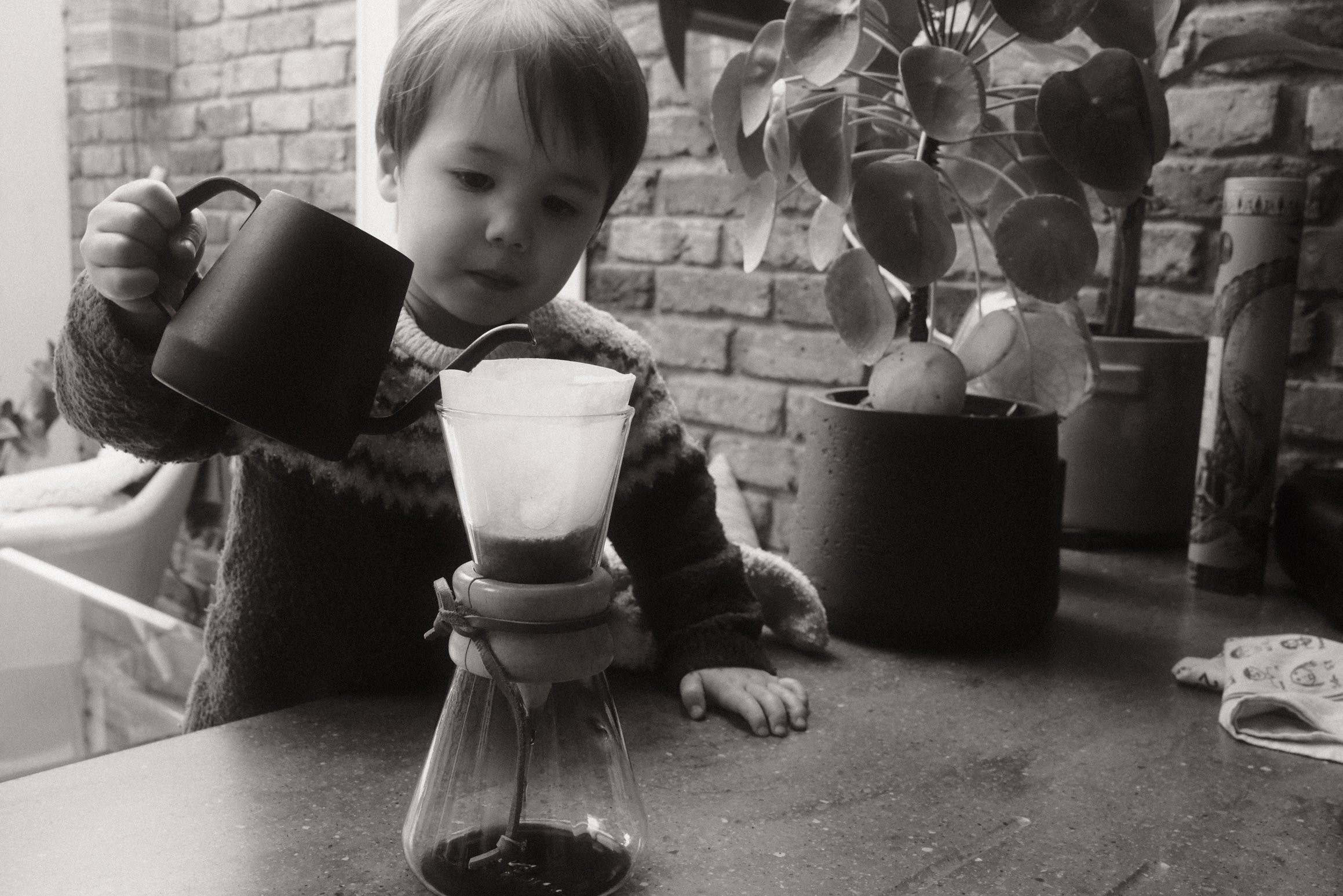 a black and white photo of a boy pouring water into a filter coffee