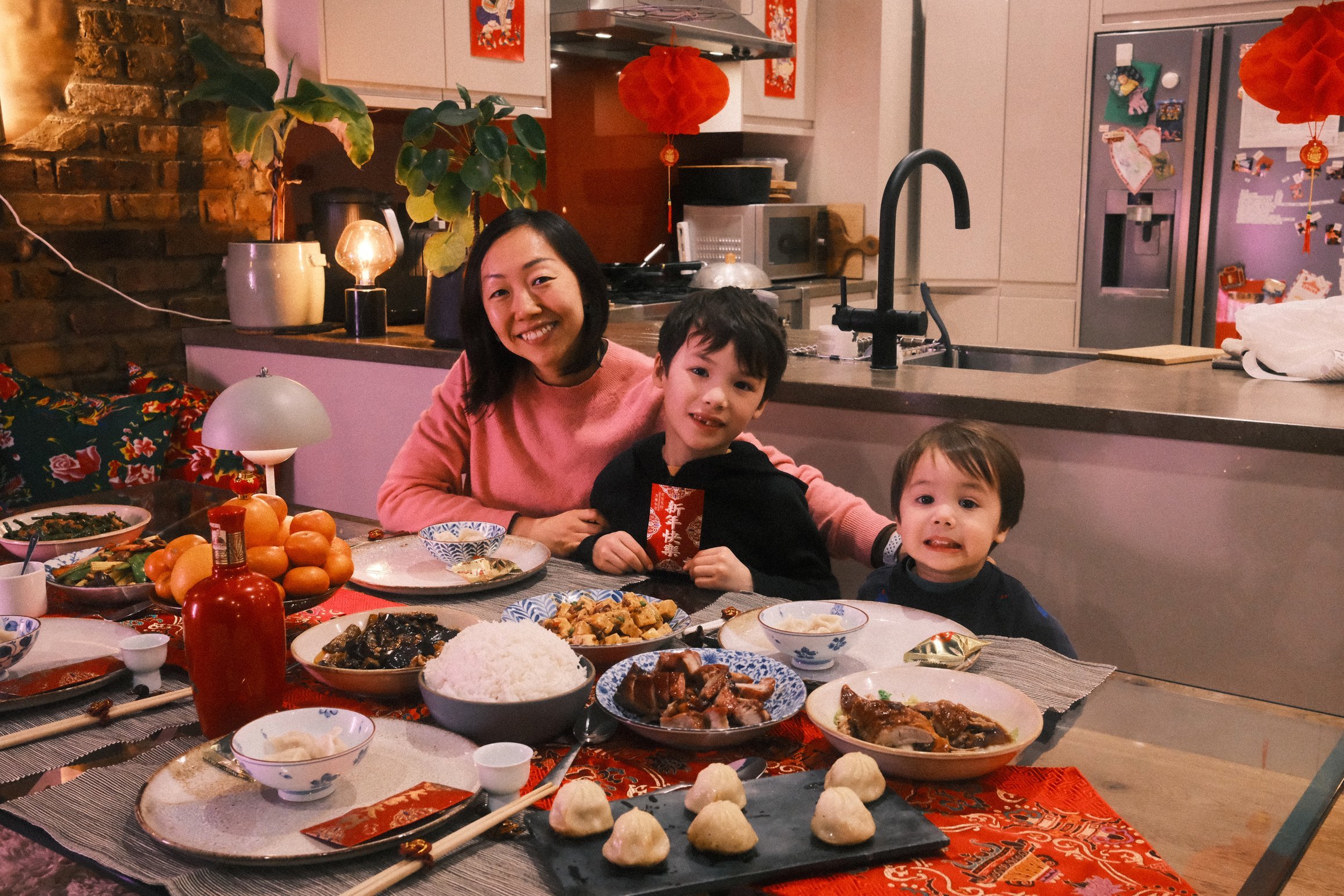 A mother and her two boys sit at a table covered in food. One boy is holding a red packet. Red lanterns hang in the background.
