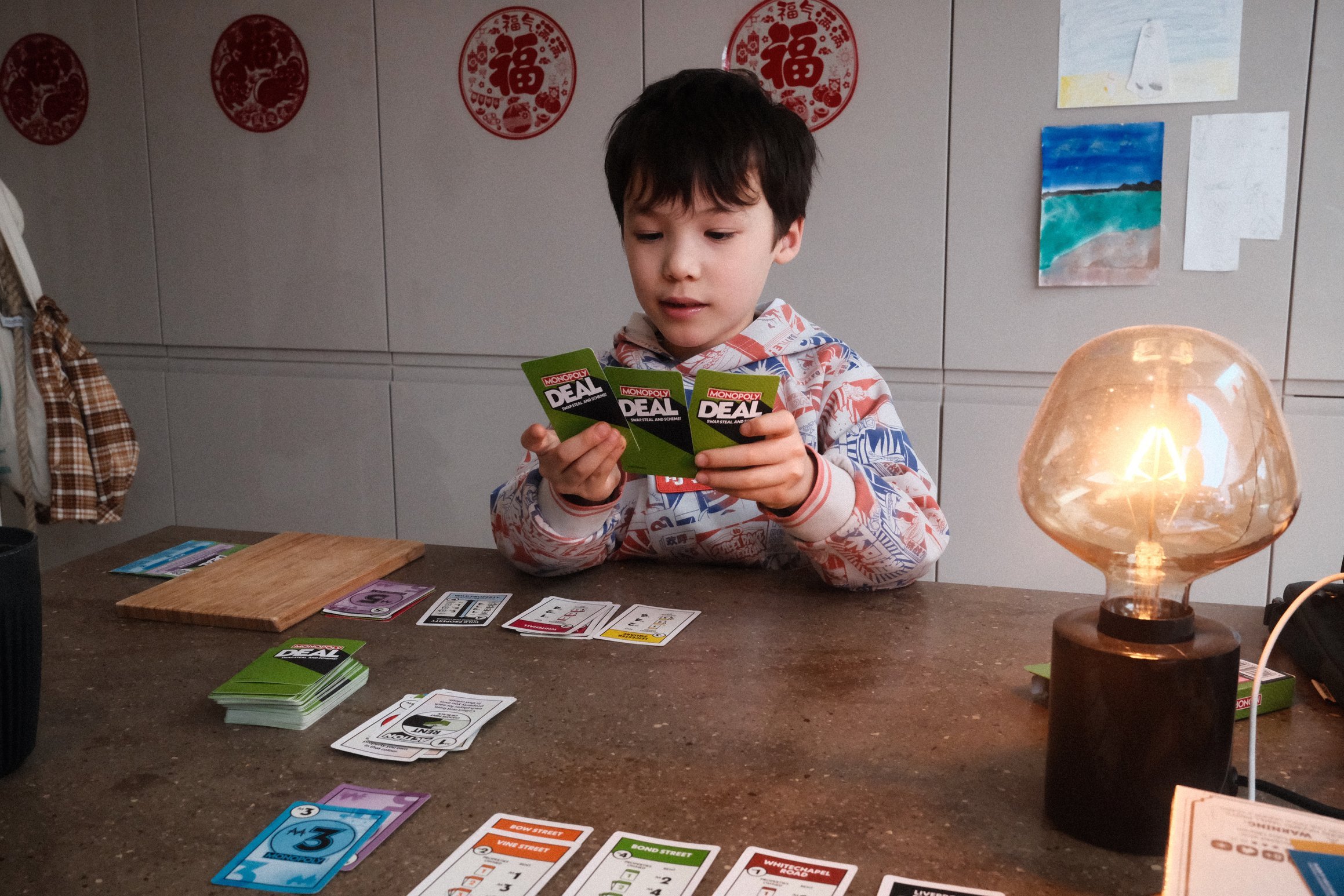 A boy holds three cards in a game of Monopoly Deal. Behind him, decorations for the Chinese New Year are mounted on the wall.