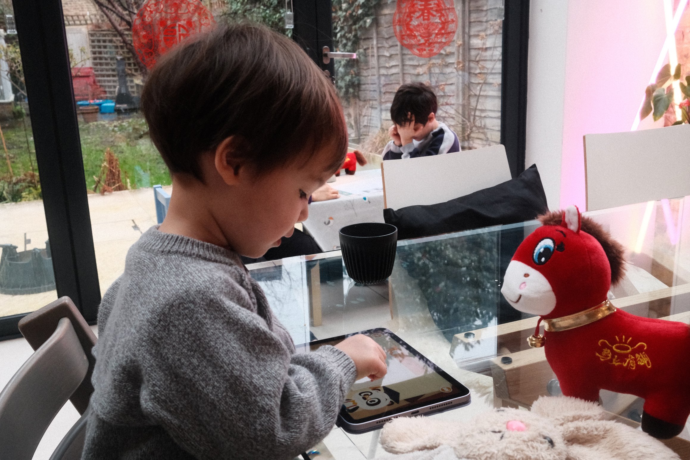 A young boy draws with his finger on an iPad, next to him a rabbit and a red horse watch him work. In the background an older boy sits at a small table, head in his hands thinking.