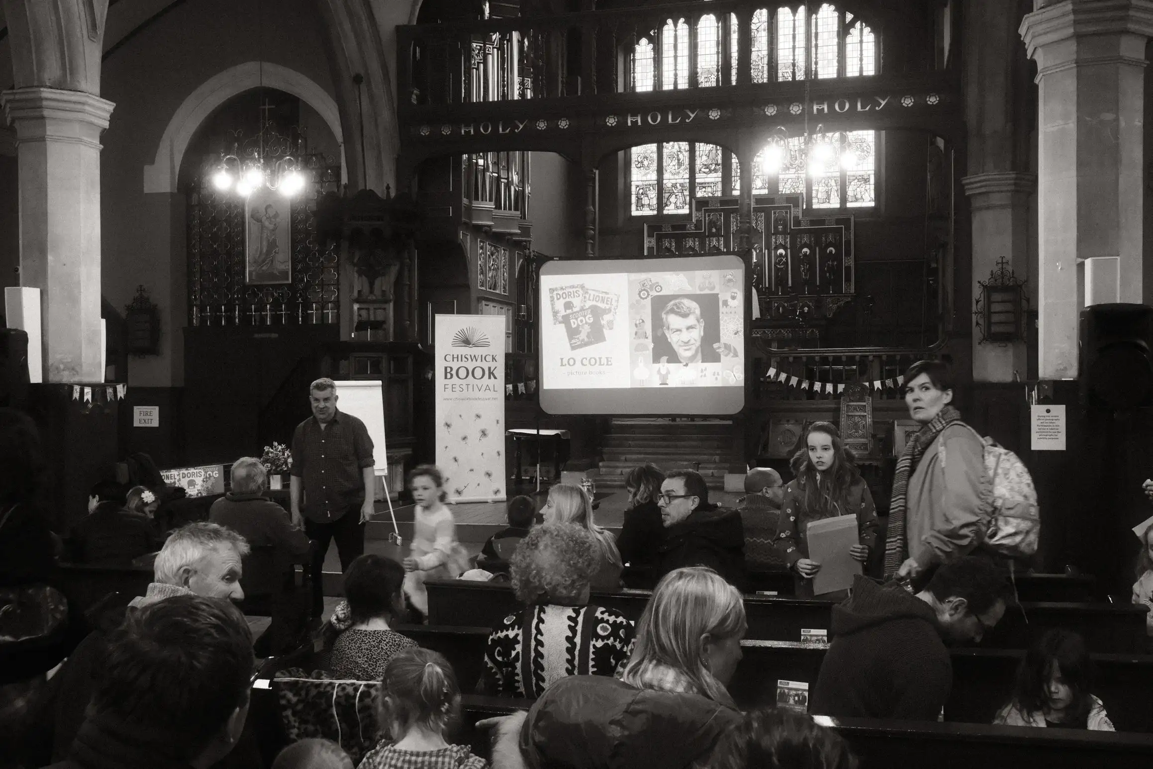 A black and white photo of Lo Cole, author, speaking at Chiswick Book Festival. The festival takes place inside a church.