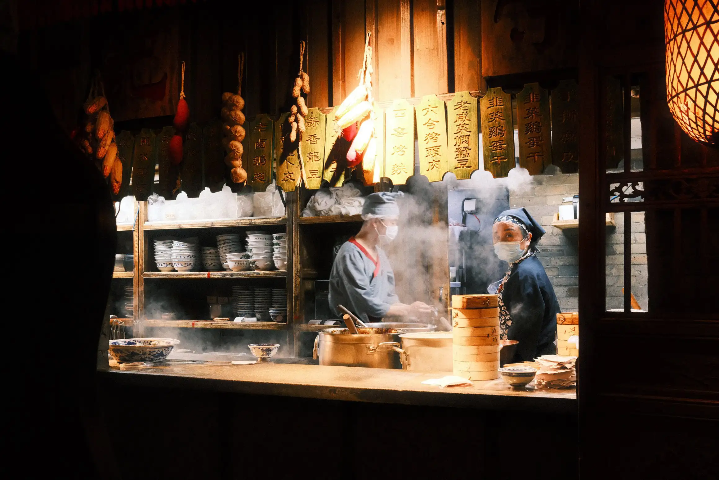 A photograph of two chefs at work in a Chinese kitchen. A bamboo steamer stands on the pass. Steam rises from a wok between the chefs.