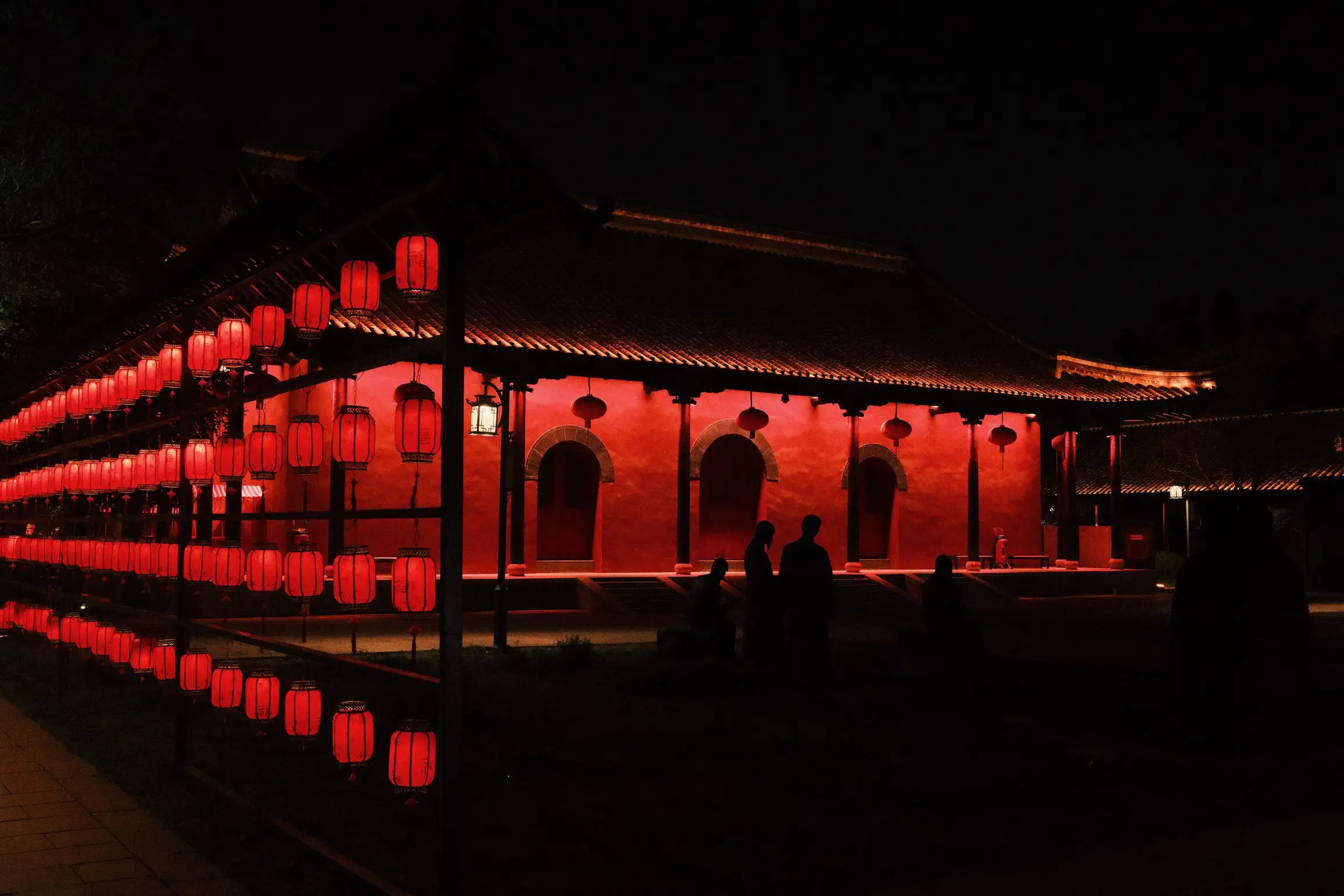 A nighttime photo of a red temple. A family silhouetted in front of the brightly lit walls. Down the left side of the photo are four rows of red lanterns stretching off into the distance.