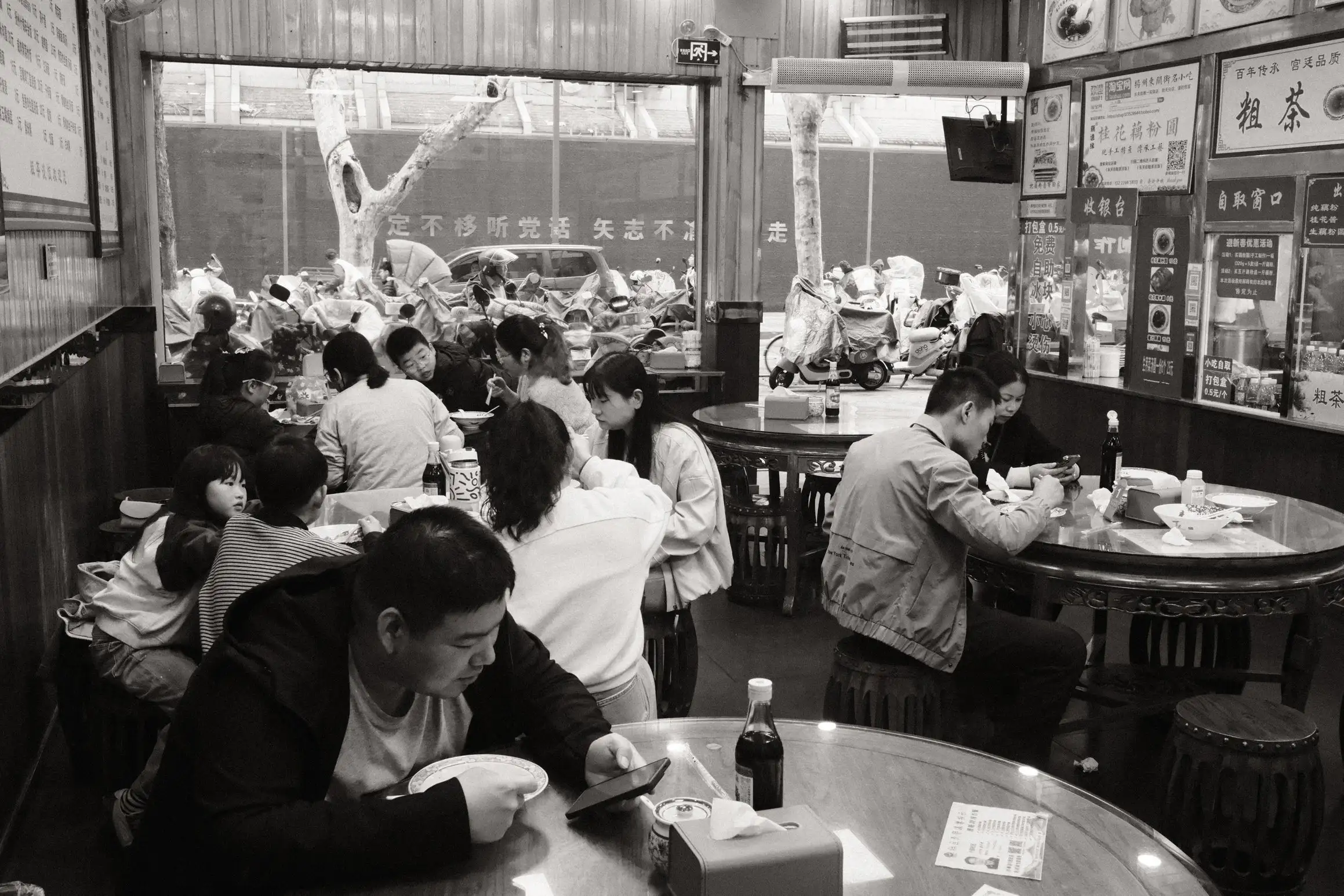 A black and white table of a Chinese restaurant looking out towards the street. In the main dining area, five round tables are occupied with people eating breakfast. The closest table has one occupant but tables further away are occupied by families.