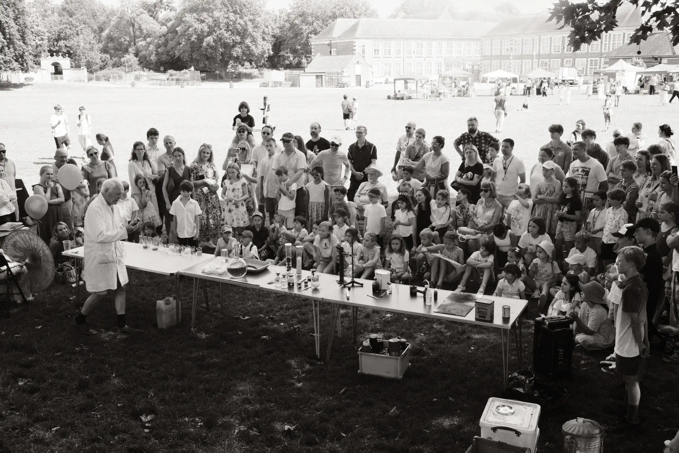 Black and white and white photo of a crowd of people facing towards a row of tables. On the tables, an array of scientific apparatus. In front of them, an old man in a white lab coat performs experiments.