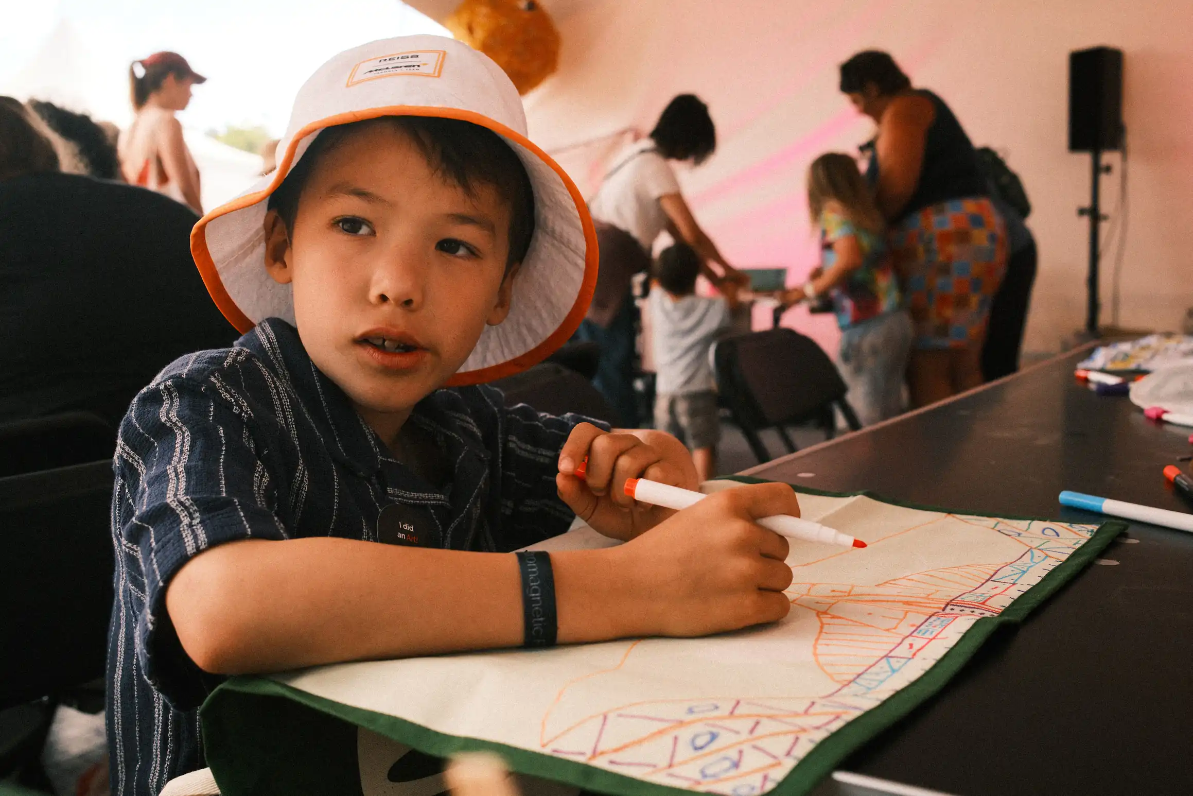 sA boy looks off to his right, in his hand a felt tip pen, in front of him an upturned tea-tail on the tea-tail he has been drawing a pattern 