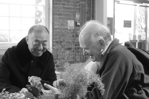Two men sit at a table smiling but they are not looking at each other. They are both looking down towards their phones. This moment of shared joy was a connection brought about through technology, translation software allowing two people to exchange ideas and experiences despite a language barrier.